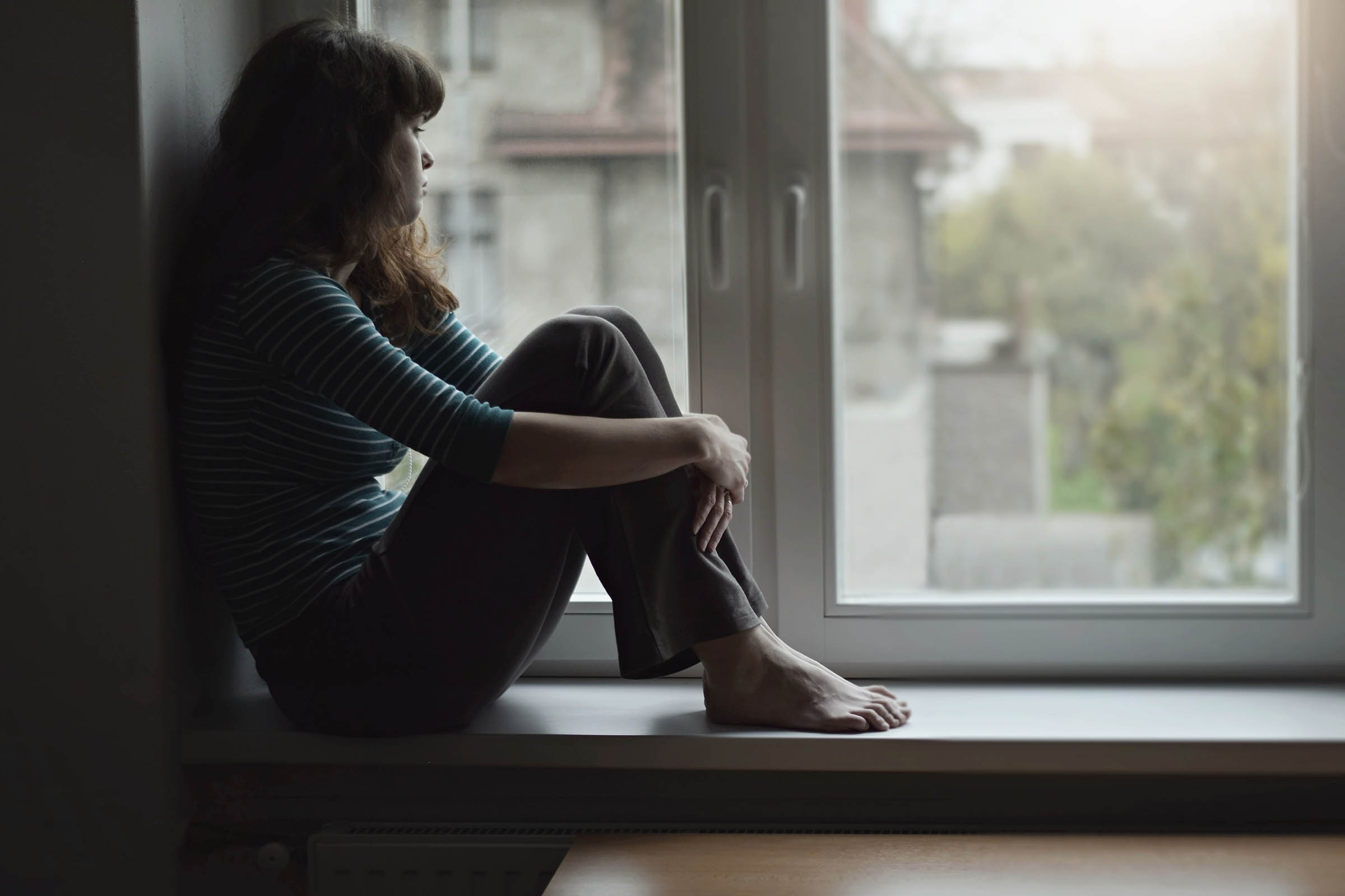 Sad young woman sitting on the window at home isolated, watching out. Coronavirus quarantine concept.
